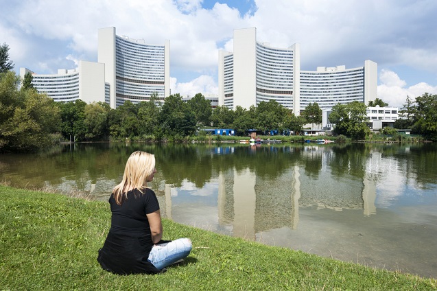 Frau sitzt vor Gewässer, im Hintergrund die Gebäude der UNO City in Wien